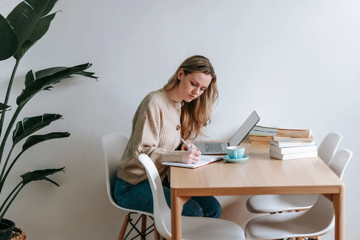 Una persona scrive a un tavolo luminoso, tra libri, appunti e laptop.